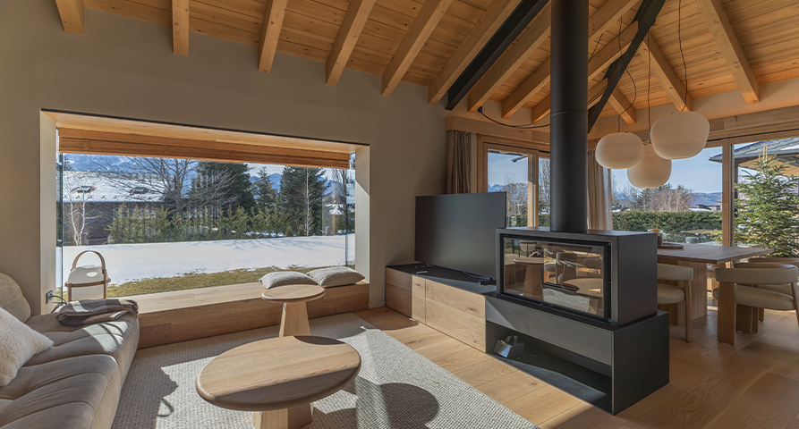 Sala d'estar de Casa L'Horitzó, amb sostre de fusta inclinat i bigues vistes, llar de foc central de disseny contemporani, sofà clar i taules baixes de fusta. Gran finestra panoràmica amb vistes a un jardí nevat i muntanyes al fons.